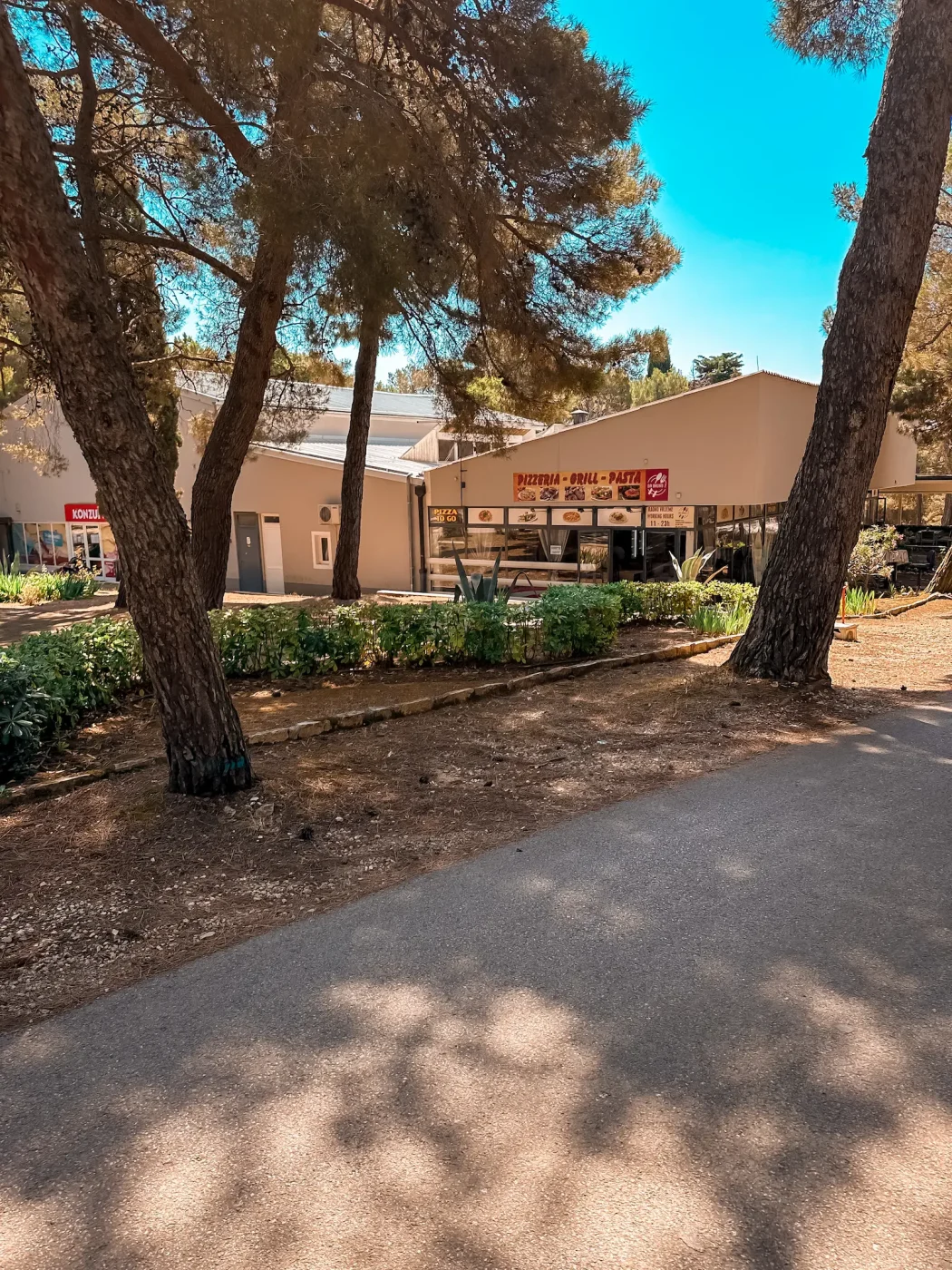 Blick auf den Konzum-Markt und die Pizzeria im schattigen Pinienwald des Camping Arena Stupice