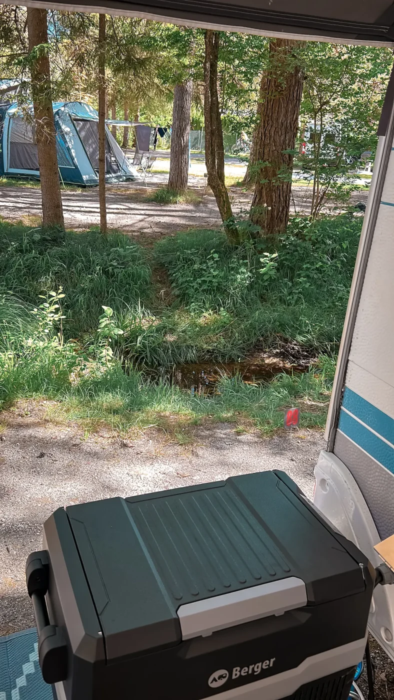 Blick vom Wohnwagen auf einen kleinen Bach, Bäume und benachbarte Stellplätze bei Strandcamping Gruber